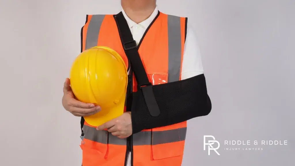 Person in an orange safety vest and arm sling holds a yellow hard hat