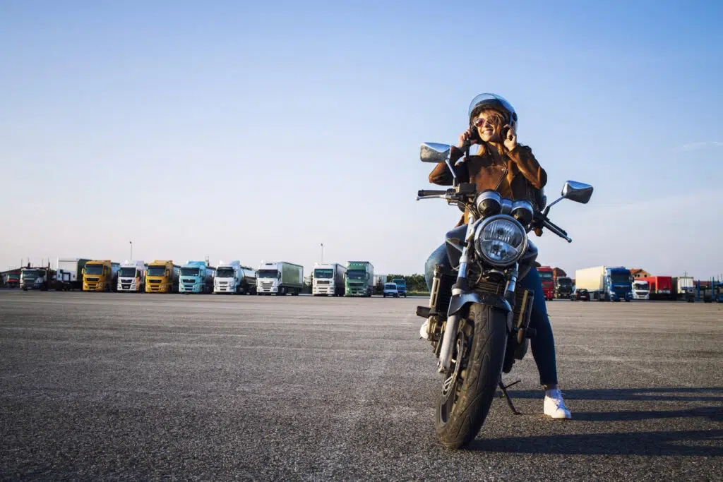 Woman taking off her helmet while on a motorcycle with semi trucks in the background