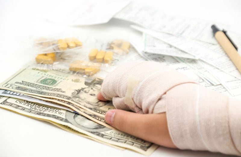 A hand in a cast receiving a stack of money, with painkillers and prescriptions in the background - Riddle & Brantley