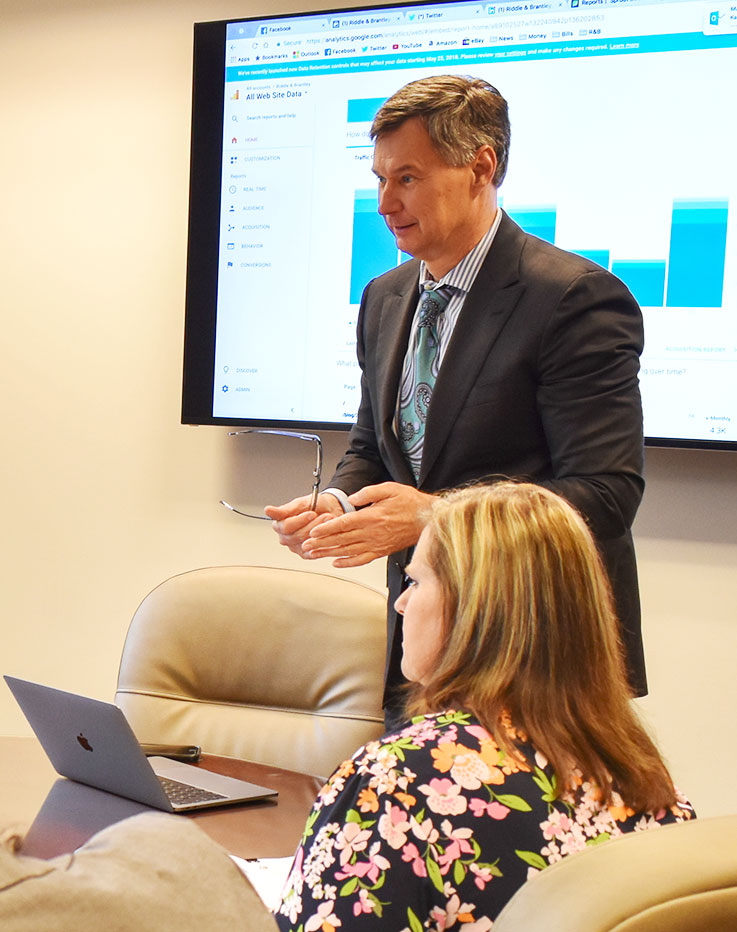 Gene Riddle in front of a projector screen showing charts and graphs