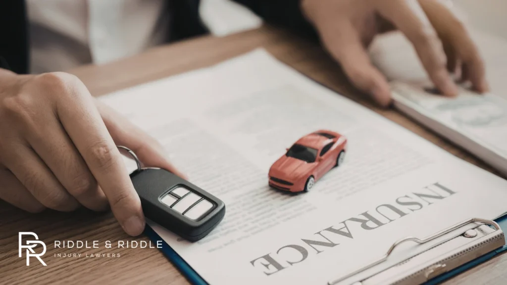 Hand holds a car key fob over an insurance document with two small toy cars nearby