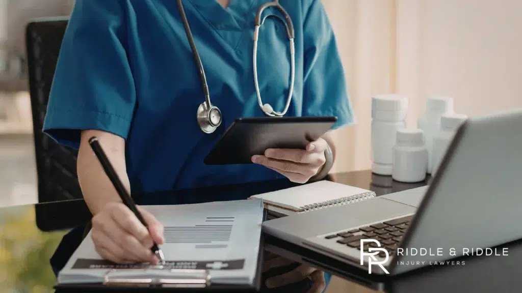 Close-up of a healthcare worker in blue scrubs filling out forms next to a laptop