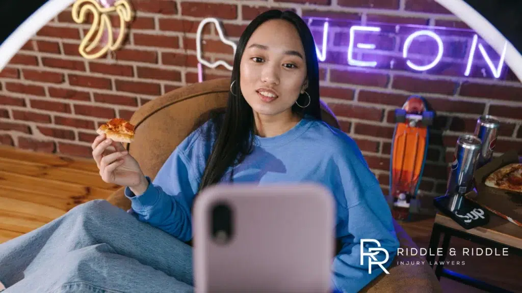 A woman eats pizza while filming a video on her smartphone in a room with neon signs