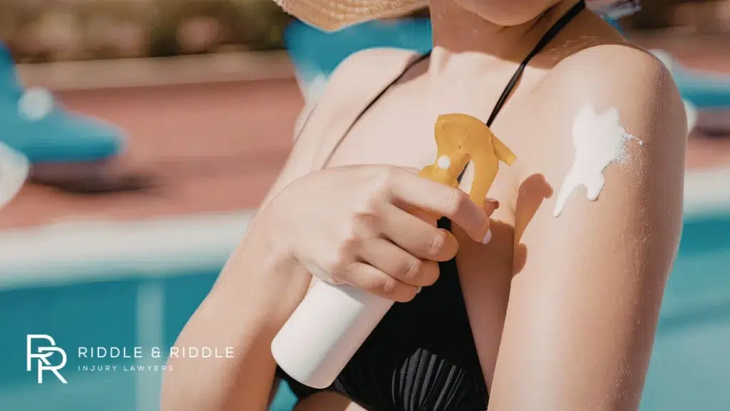 Woman in a sun hat sprays white sunscreen onto her shoulder by a swimming pool