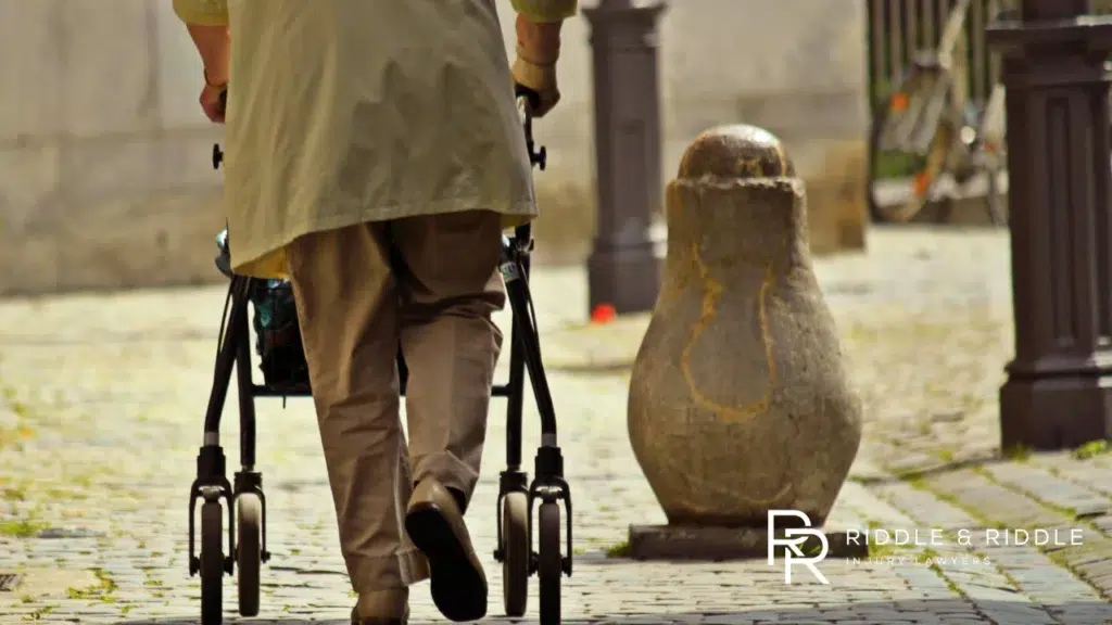Person viewed from behind using a four-wheeled walker to navigate a paved outdoor path