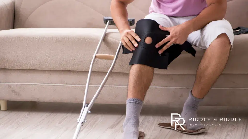 Person sitting on a sofa wears a black knee brace with a crutch leaning nearby