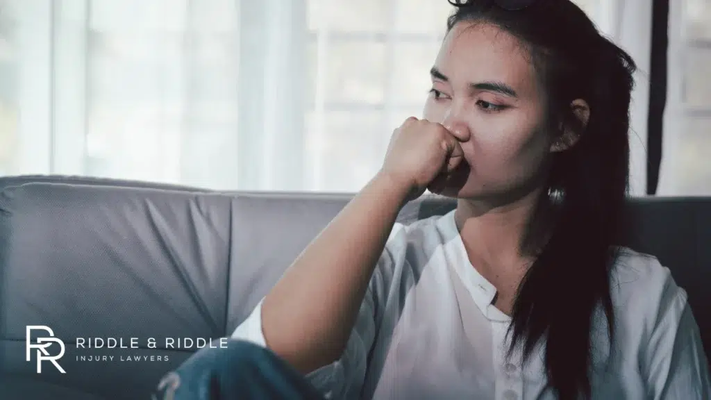 Young woman sits on a sofa looking thoughtfully out a window, resting her chin on her hand
