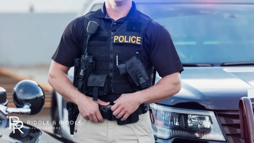 Police officer in uniform and tactical vest stands in front of a patrol car