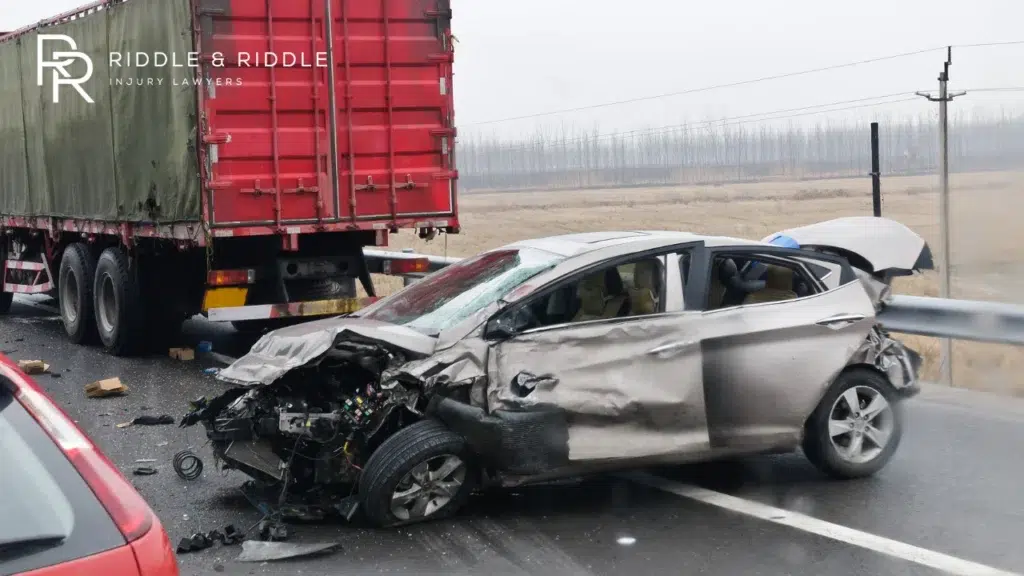 Silver car with a completely destroyed front end next to a large red shipping truck
