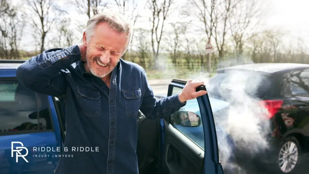 An older man rubs his neck in pain while standing next to a smoking car after an accident