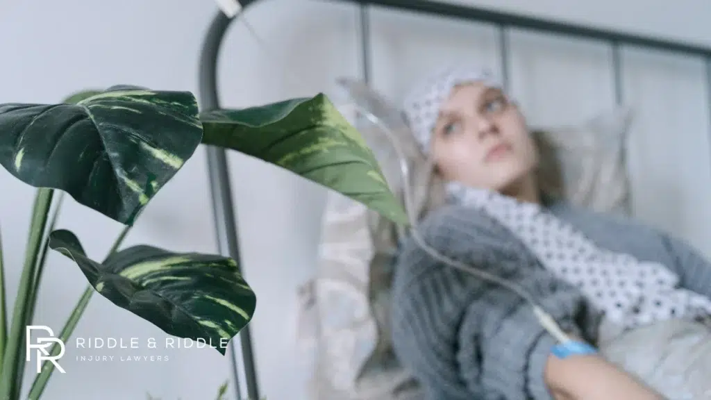young woman in a hospital gown lies in bed with an IV drip and a plant in the foreground