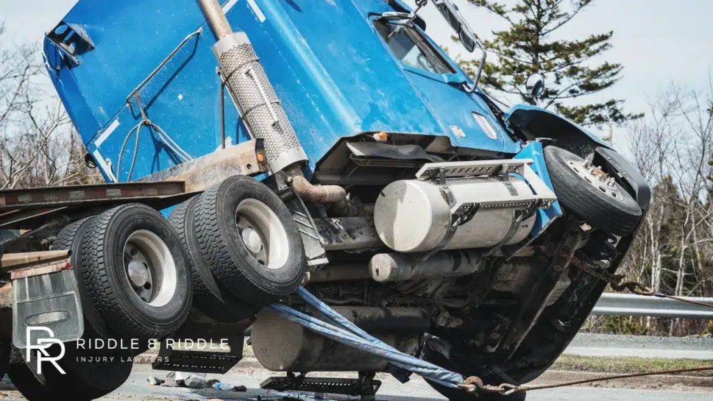 Large blue semi-truck lying completely on its side off the edge of a paved road