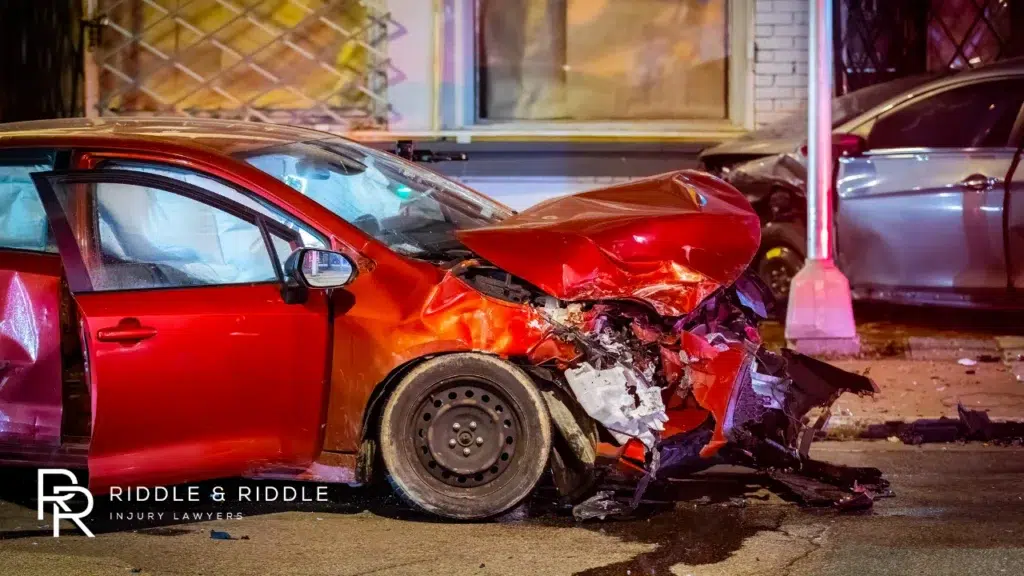 Red car with severe front-end damage parked on a city street at night