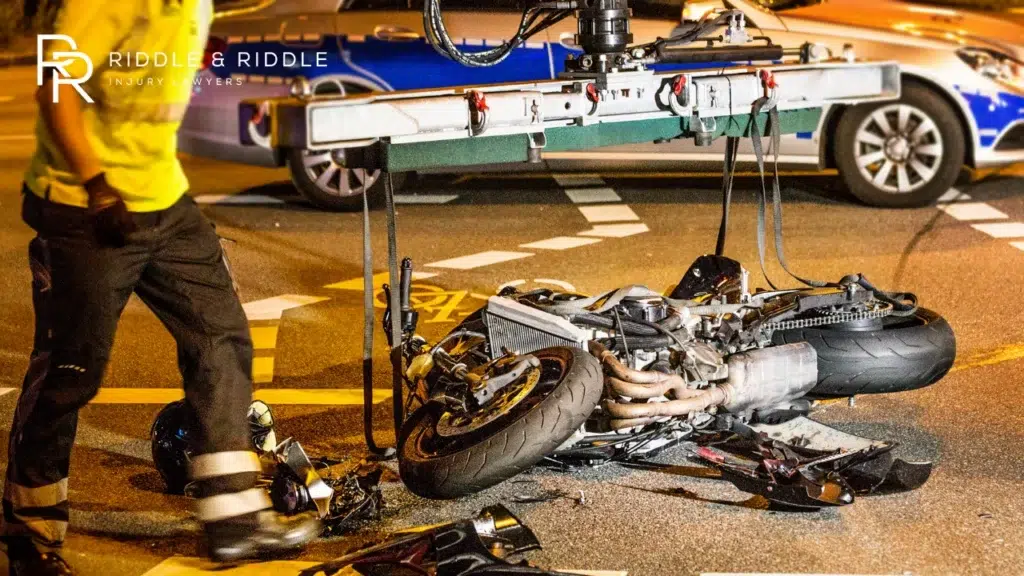 Wrecked motorcycle lies on the pavement at night with a person in a yellow shirt nearby