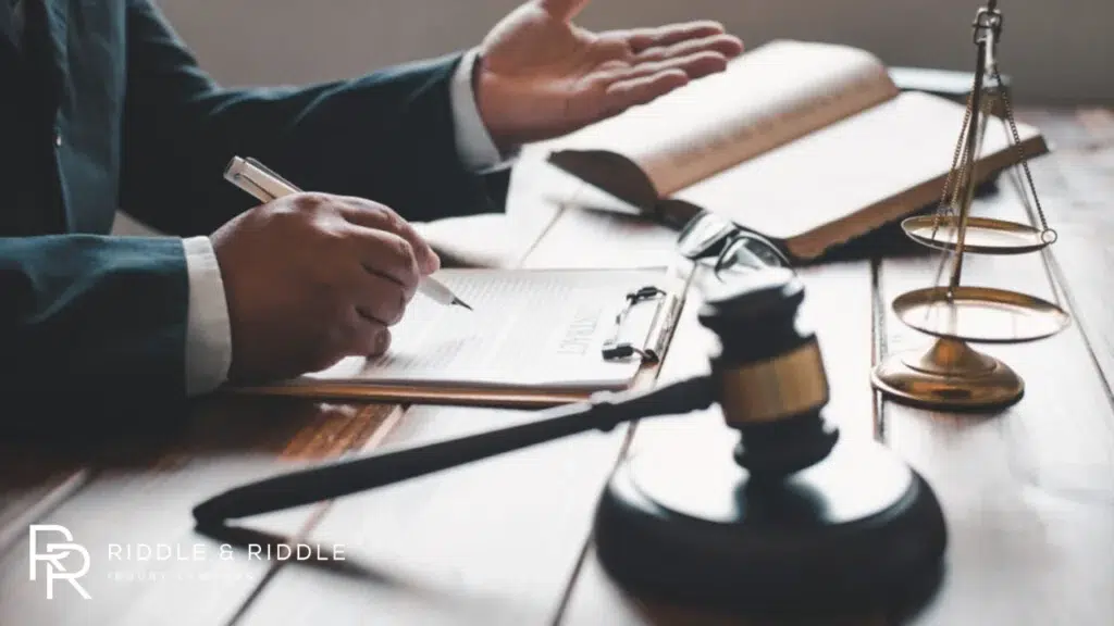 A legal professional is writing on documents at a desk featuring an open book and a set of scales of justice.